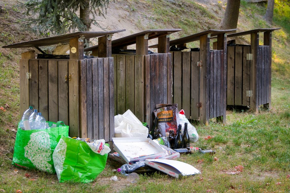 A row of four small, weathered wooden outdoor compost or waste bins with sloped dark brown roofs, positioned on a grassy area beside a wooded slope. The bins are made of vertical wooden planks, some s