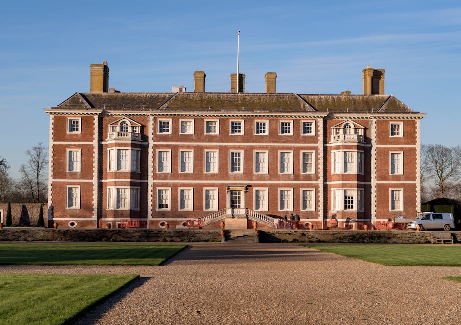 A large, historic brick building with four symmetrical stories, set against a clear blue sky. The facade features numerous white-framed sash windows, with decorative stone or plaster accents around so